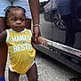 Ace is registered to the contest to win money with this photo: toddler, child, yellow_onesie, white_shoes, braided_hair, adult_hand, car, reflection, pavement, mismatched_socks, sandals, outdoor, walking, support, person, road, casual_clothing, necklace, summer, baby