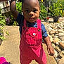 toddler, child, outdoor, sunlight, red_overalls, striped_clothing, blue_shirt, pigtails, gold_chain, holding_hand, garden, plants, rocks, sidewalk, shoes, curious, portrait, person, summer, daylight