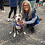 woman, dog, smiling, glasses, harness, asphalt, people, outdoor, event, casual, sneakers, blue_clothing, gray_dog, white_dog, leash, social_gathering, standing, crouching, happy, friendly