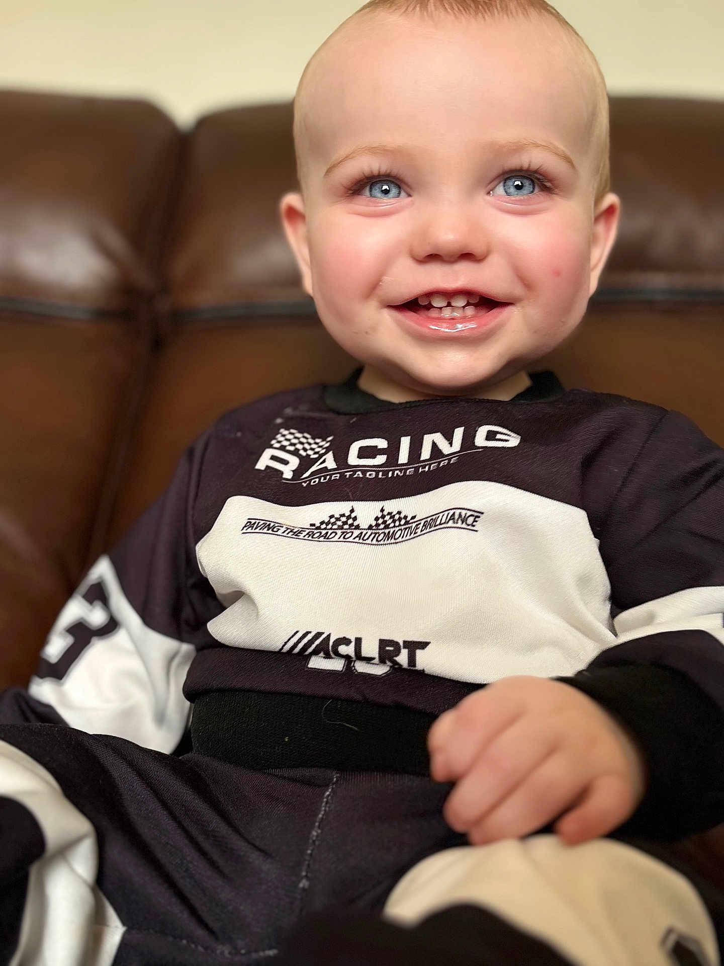 Harry is registered to the contest to win money with this photo: toddler, child, smiling, blue_eyes, racing_outfit, sitting, couch, leather, happy, portrait, indoor, clothing, cute, baby, person, young_child, face, hand, casual, closeup