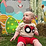 baby, child, grass, red_shorts, beige_shirt, tractor_design, cartoon_bunny, colorful_backdrop, flowers, sunflower, hay_bales, outdoor, playful, cute, portrait, sitting, curious, blue_eyes, soft_light, decorations