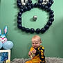 Harry is registered to the contest to win money with this photo: baby, child, balloons, easter, plush_rug, toy_duck, yellow_outfit, foil_wrapper, balloon_art, green_wall, indoor, celebration, decorations, festive, sitting, unwrapping, cute, holiday, happy, person