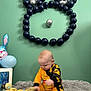 Harry is registered to the contest to win money with this photo: baby, child, plush_toy, balloon, yellow_clothing, black_clothing, soft_rug, green_wall, decorations, stuffed_duck, stuffed_animal, indoor, playful, cute, baby_sitting, balloon_art, toy, candid, infant, happy