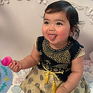 Aisha is registered to the contest to win money with this photo: baby, child, tongue_out, playful, black_hair, cute, dress, patterned_dress, gold_embroidery, toy, pink_toy, sitting, blanket, colorful_blanket, indoor, portrait, happy, chubby_cheeks, face, person