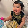 baby, child, tongue_out, playful, black_hair, cute, dress, patterned_dress, gold_embroidery, toy, pink_toy, sitting, blanket, colorful_blanket, indoor, portrait, happy, chubby_cheeks, face, person