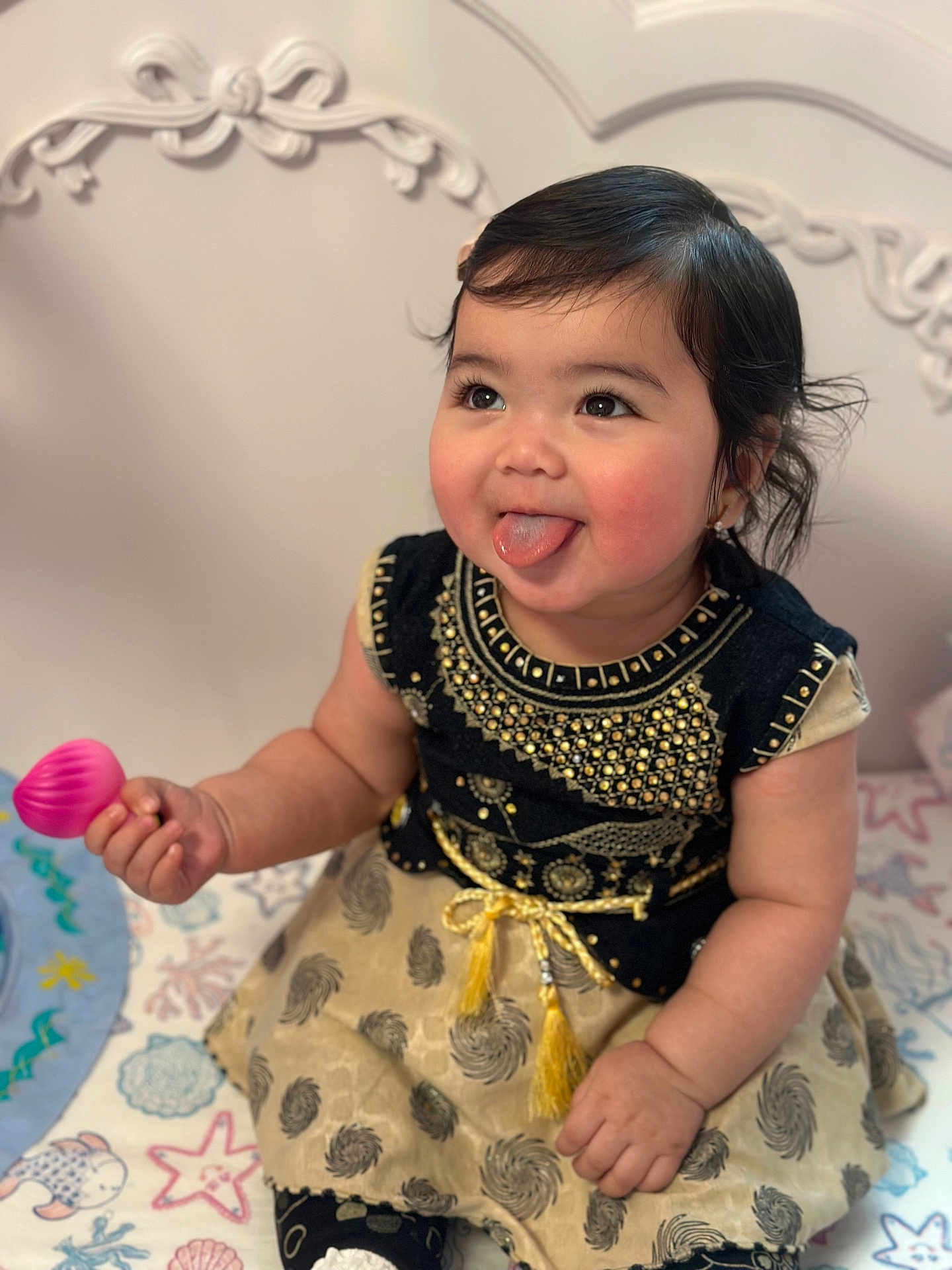 Aisha is registered to the contest to win money with this photo: baby, child, tongue_out, playful, black_hair, cute, dress, patterned_dress, gold_embroidery, toy, pink_toy, sitting, blanket, colorful_blanket, indoor, portrait, happy, chubby_cheeks, face, person