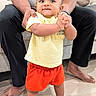 Viraj Reddy joined the competition — help win amazing prizes! toddler, baby, child, standing, indoor, adult, hands, clothing, yellow_shirt, orange_shorts, barefoot, wooden_floor, couch, casual_wear, person, supporting, home, portrait, curious, bright_colors