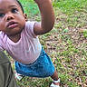 toddler, child, outdoor, grass, curious, denim_skirt, pink_shirt, white_sandals, reaching_hand, close_up, nature, young_child, exploration, casual_clothing, summer, person, portrait, grass_field, daylight, cute