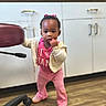 toddler, child, indoor, standing, pink_clothing, jacket, shoes, wooden_floor, cabinet, chair, curly_hair, bracelet, expression, furniture, support, young_child, person, flooring, holding, casual_wear