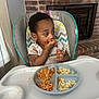 child, baby, high_chair, food, spaghetti, scrambled_eggs, messy, eating, indoor, plate, feeding, person, face, clothing, curtain, fireplace, brick_wall, table, hand, meal