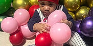 Elijah is registered to the contest to win money with this photo: baby, child, balloons, red_beanie, striped_outfit, indoor, carpet, colorful, party, celebration, sitting, curious, holding, pink_balloons, red_balloons, green_balloons, gold_balloons, purple_balloons, cute, infant