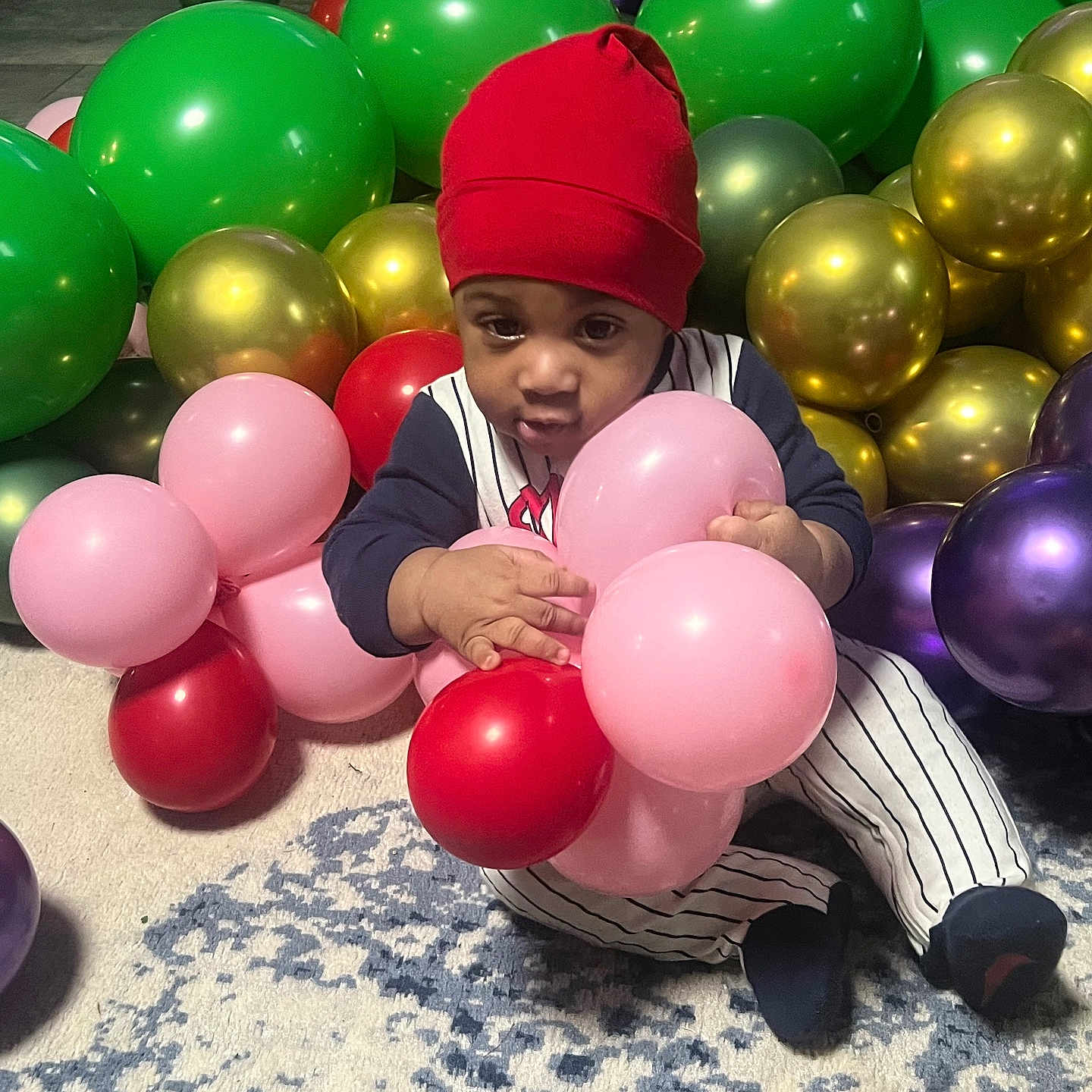 Elijah is registered to the contest to win money with this photo: baby, balloons, carpet, celebration, child, colorful, curious, cute, gold_balloons, green_balloons, holding, indoor, infant, party, pink_balloons, purple_balloons, red_balloons, red_beanie, sitting, striped_outfit