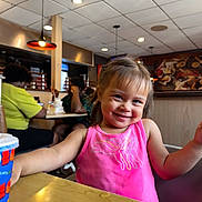 Aylani joined the competition — help win amazing prizes! toddler, child, smile, pink_shirt, restaurant, table, cup, indoor, people, background, happy, face, hairstyle, seat, lighting, casual, dining, fun, person, portrait