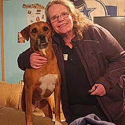 Molly Lou is registered to the contest to win money with this photo: blanket, brown_dog, casual_clothing, couch, cozy, dog, flag, furniture, glasses, hat, home, hot_wheels, indoor, person, pet, smiling, television, wall_decor, white_patch, woman