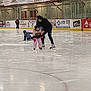 child, adult, ice_rink, ice_skating, skates, helmet, winter_clothing, frozen_surface, advertisements, sports, falling, helping, indoor, gloves, jacket, pants, rink_board, people, activity