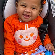 Kalena is registered to the contest to win money with this photo: baby, child, car_seat, orange_clothing, bow, smiling, toy, patterned_pants, infant, cute, happy, portrait, indoor, seatbelt, playful, closeup, skin, hair, cheeks, hand