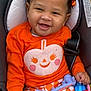 baby, child, car_seat, orange_clothing, bow, smiling, toy, patterned_pants, infant, cute, happy, portrait, indoor, seatbelt, playful, closeup, skin, hair, cheeks, hand