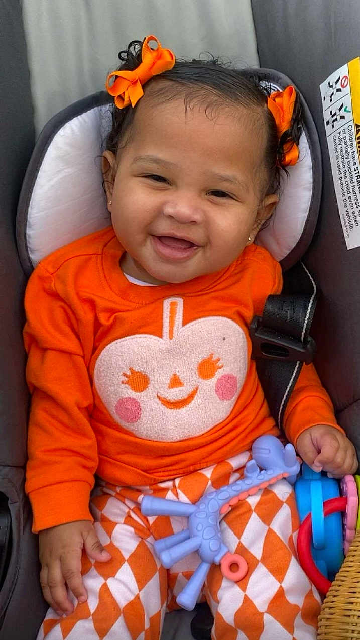 Kalena is registered to the contest to win money with this photo: baby, child, car_seat, orange_clothing, bow, smiling, toy, patterned_pants, infant, cute, happy, portrait, indoor, seatbelt, playful, closeup, skin, hair, cheeks, hand