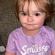 Elayna is registered to the contest to win money with this photo: child, toddler, smile, purple_shirt, text_on_clothing, sitting, brown_eyes, hair, indoors, couch, fabric, cute, person, portrait, cozy, face, expression, casual_clothing, young, happy