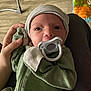 Gunner is registered to the contest to win money with this photo: baby, infant, newborn, pacifier, hat, hand, onesie, green_clothing, face, eyes, lap, indoor, wood_floor, toy, baby_bouncer, parent_hand, portrait, clothing_zipper, sitting, screenshot_overlay