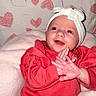 baby, infant, child, red_clothing, headband, bow, clapping_hands, smiling, happy, heart_pattern, blanket, indoors, cute, portrait, young_child, warm_clothing, pink_blanket, background_pattern, seated, playful