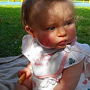 Lou a rejoint le concours — aidez-le/la à gagner de superbes lots ! baby, bib, blanket, child, clothing, curly_hair, daylight, expression, face, grass, outdoor, park, play_equipment, playground, ponytail, shadow, sitting, snack, sunlight, toddler