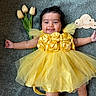 baby, yellow_dress, tulips, flower, smile, infant, child, portrait, happy, laying_down, cute, roses, fabric, skin, feet, hand, wooden_sign, celebration, indoors, person