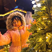 Ariana joined the competition — help win amazing prizes!  festive, child, christmas_decorations, christmas_tree, face, fur_hood, hair, holiday, indoor, lights, orange_clothing, ornaments, parka, portrait, reaching, smiling, standing, warm_lighting, winter, young_person