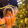  festive, child, christmas_decorations, christmas_tree, face, fur_hood, hair, holiday, indoor, lights, orange_clothing, ornaments, parka, portrait, reaching, smiling, standing, warm_lighting, winter, young_person