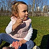 child, toddler, grass, flower, outdoor, sunny, park, puffy_vest, hair_clip, blue_sky, trees, hand, shoes, curious, sitting, nature, daylight, casual_clothing, young_child, playful