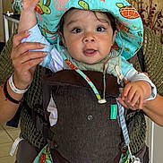 David Orro joined the competition — help win amazing prizes! baby, child, hat, colorful, turtle_pattern, carrier, indoor, person, hand, face, curious, cute, clothing, adult_hand, infant, smiling, holding, portrait, closeup, playful