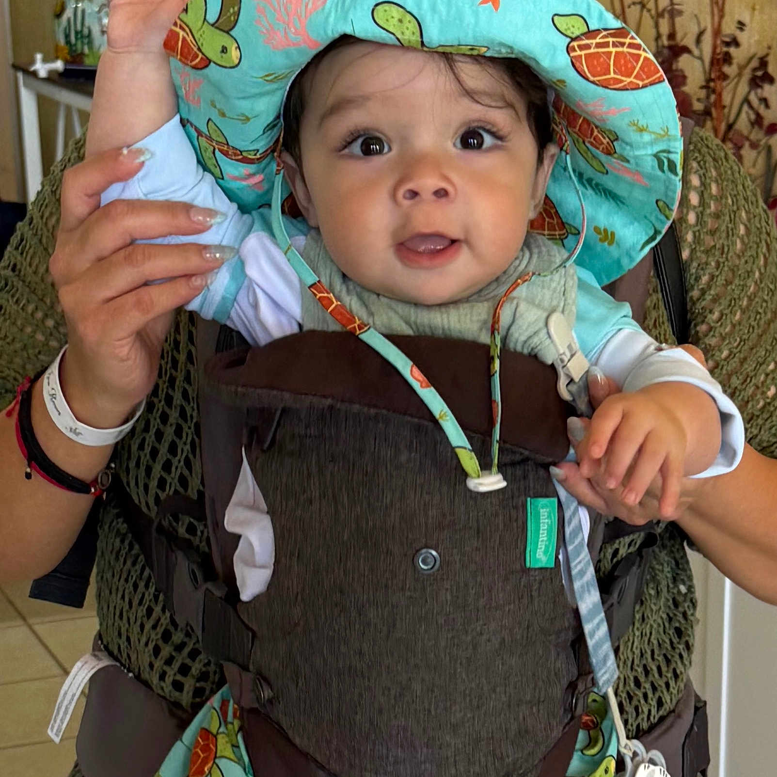 David Orro joined the competition — help win amazing prizes! baby, child, hat, colorful, turtle_pattern, carrier, indoor, person, hand, face, curious, cute, clothing, adult_hand, infant, smiling, holding, portrait, closeup, playful