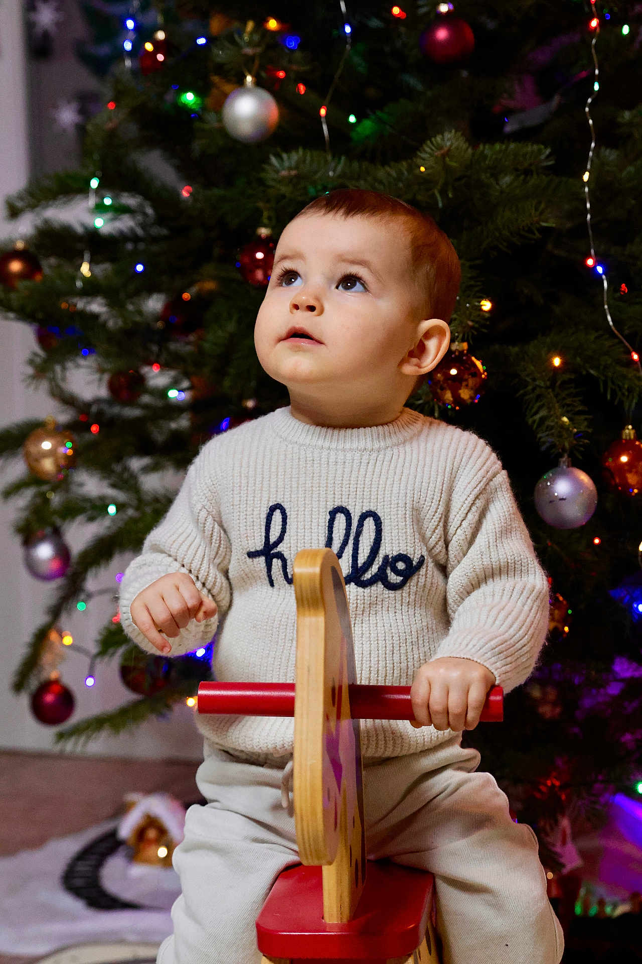Mélio a rejoint le concours — aidez-le/la à gagner de superbes lots ! toddler, child, sweater, hello, rocking_horse, toy, christmas_tree, ornaments, lights, holiday, indoor, curious, gazing, festive, decorations, seated, play, winter_clothing, cute, young_child