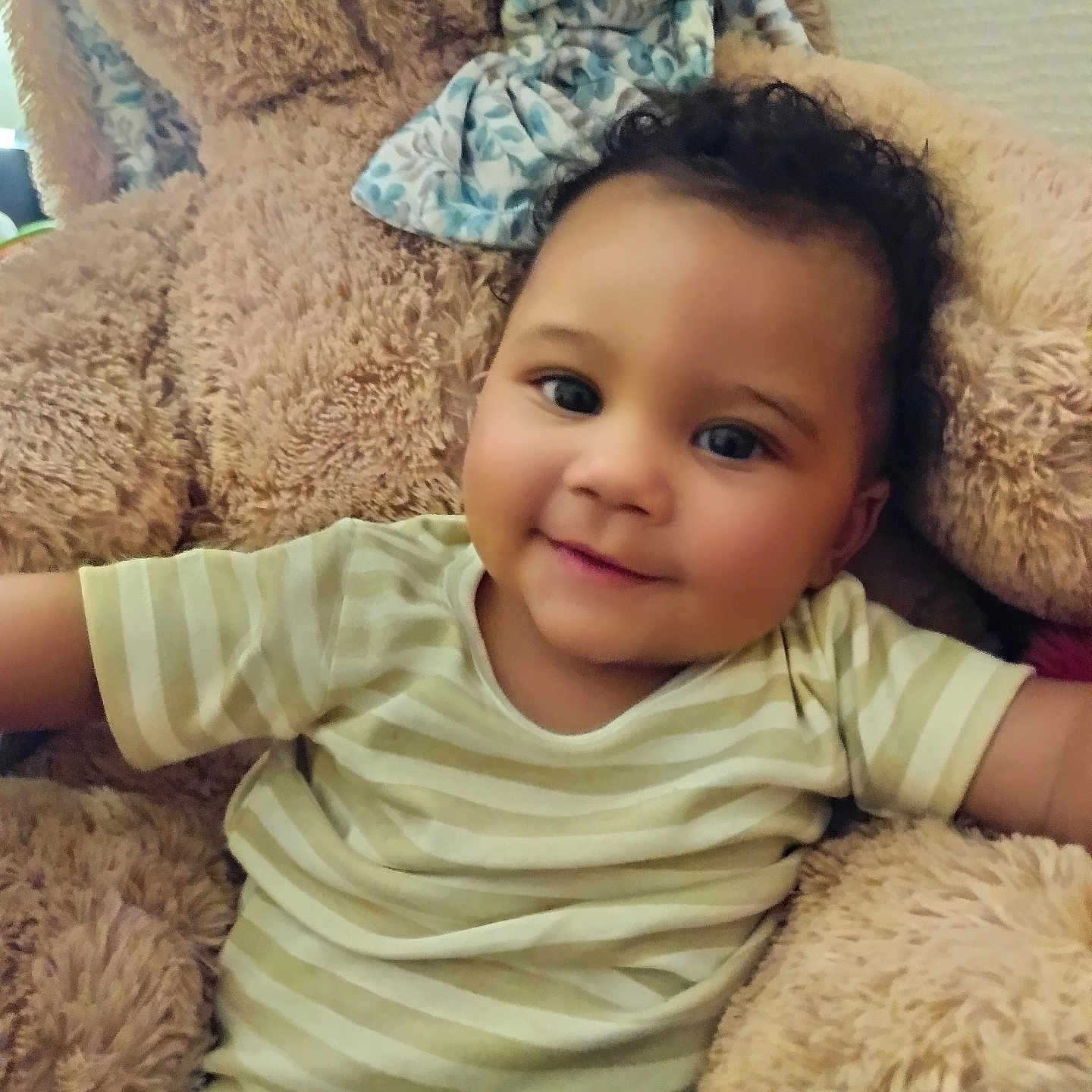 William participe au concours pour gagner de l'argent avec cette photo : baby, bow, child, cozy, curly_hair, cushion, cute, furniture, happy, indoors, person, plush, portrait, relaxing, smile, soft_toy, striped_shirt, teddy_bear, toddler, warm_lighting