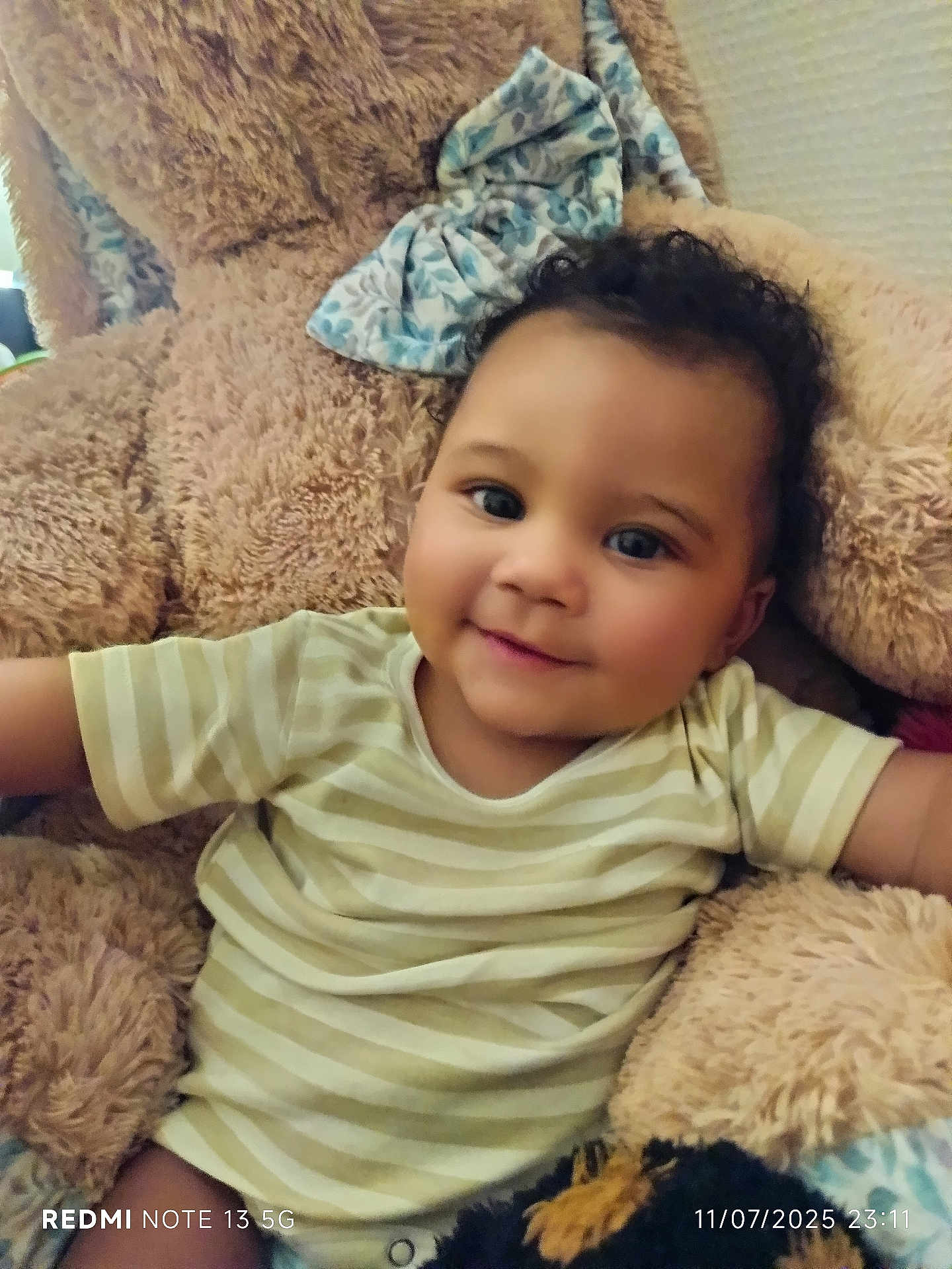 William participe au concours pour gagner de l'argent avec cette photo : toddler, child, smile, curly_hair, striped_shirt, plush, teddy_bear, bow, indoors, cozy, soft_toy, happy, portrait, baby, person, cute, furniture, cushion, relaxing, warm_lighting