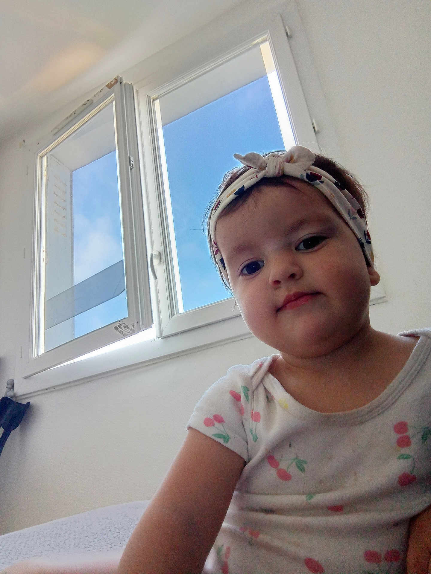 Amina participe au concours pour gagner de l'argent avec cette photo : toddler, child, indoor, window, headband, shirt, cherry_pattern, face, person, portrait, blue_sky, light, curious, expression, casual_clothing, young_child, natural_light, white_wall, home, seated