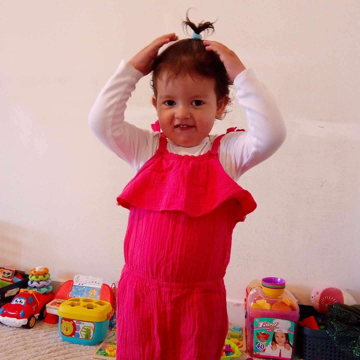 Amina is registered to the contest to win money with this photo: toddler, child, pink_romper, toy, carpet, indoor, playtime, smiling, hair_tie, colorful, room, young_child, person, cute, happy, casual, standing, fun, messy, playroom