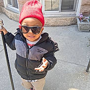 Tyrone joined the competition — help win amazing prizes! child, boy, red_beanie, sunglasses, jacket, hooded_jacket, walking, sidewalk, urban, window, stone_wall, potted_plant, casual_clothing, smiling, outdoor, daylight, person, street_style, footwear, confident