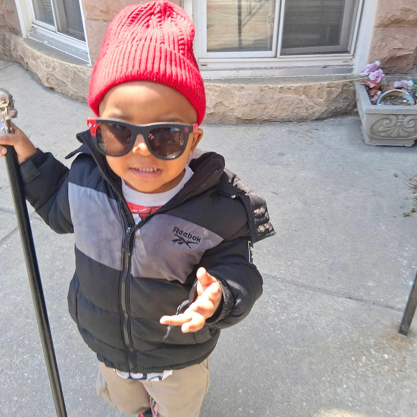 Tyrone joined the competition — help win amazing prizes! child, boy, red_beanie, sunglasses, jacket, hooded_jacket, walking, sidewalk, urban, window, stone_wall, potted_plant, casual_clothing, smiling, outdoor, daylight, person, street_style, footwear, confident