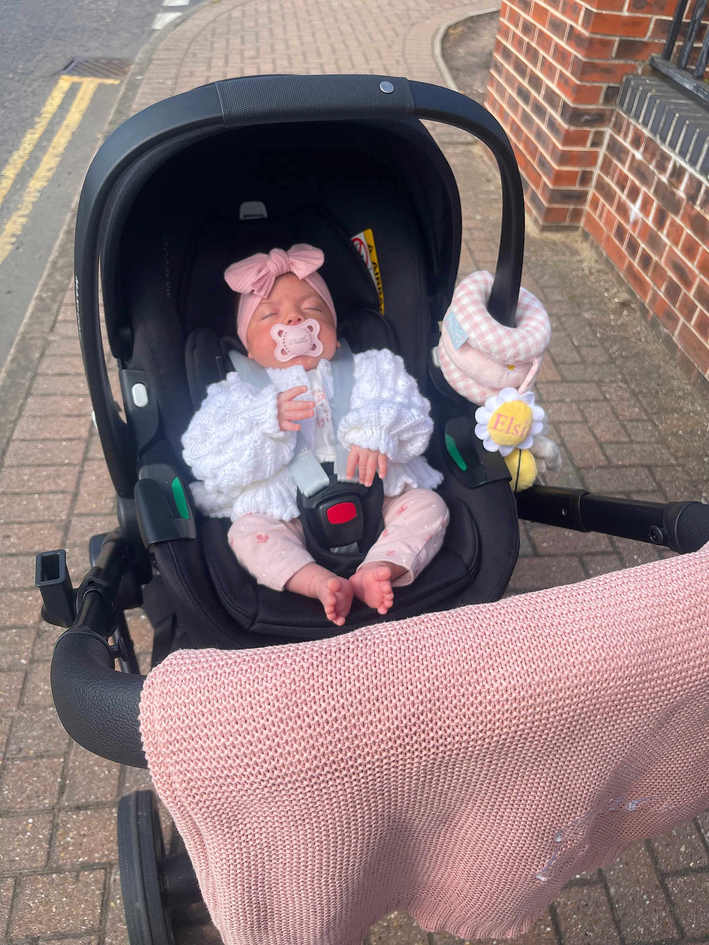Elsie is registered to the contest to win money with this photo: baby, stroller, pacifier, blanket, headband, toy, sidewalk, pavement, brick_wall, clothing, infant, person, outdoor, sleeping, cute, pink, knitwear, sitting, car_seat, child