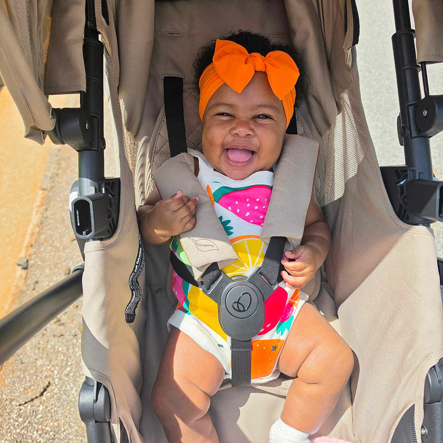 Kaci-Rose is registered to the contest to win money with this photo: baby, child, stroller, orange_bow, headband, colorful_clothing, fruit_pattern, happy, smiling, tongue_out, sitting, white_socks, sunlight, outdoor, infant, cute, seatbelt, fabric, wheels, person