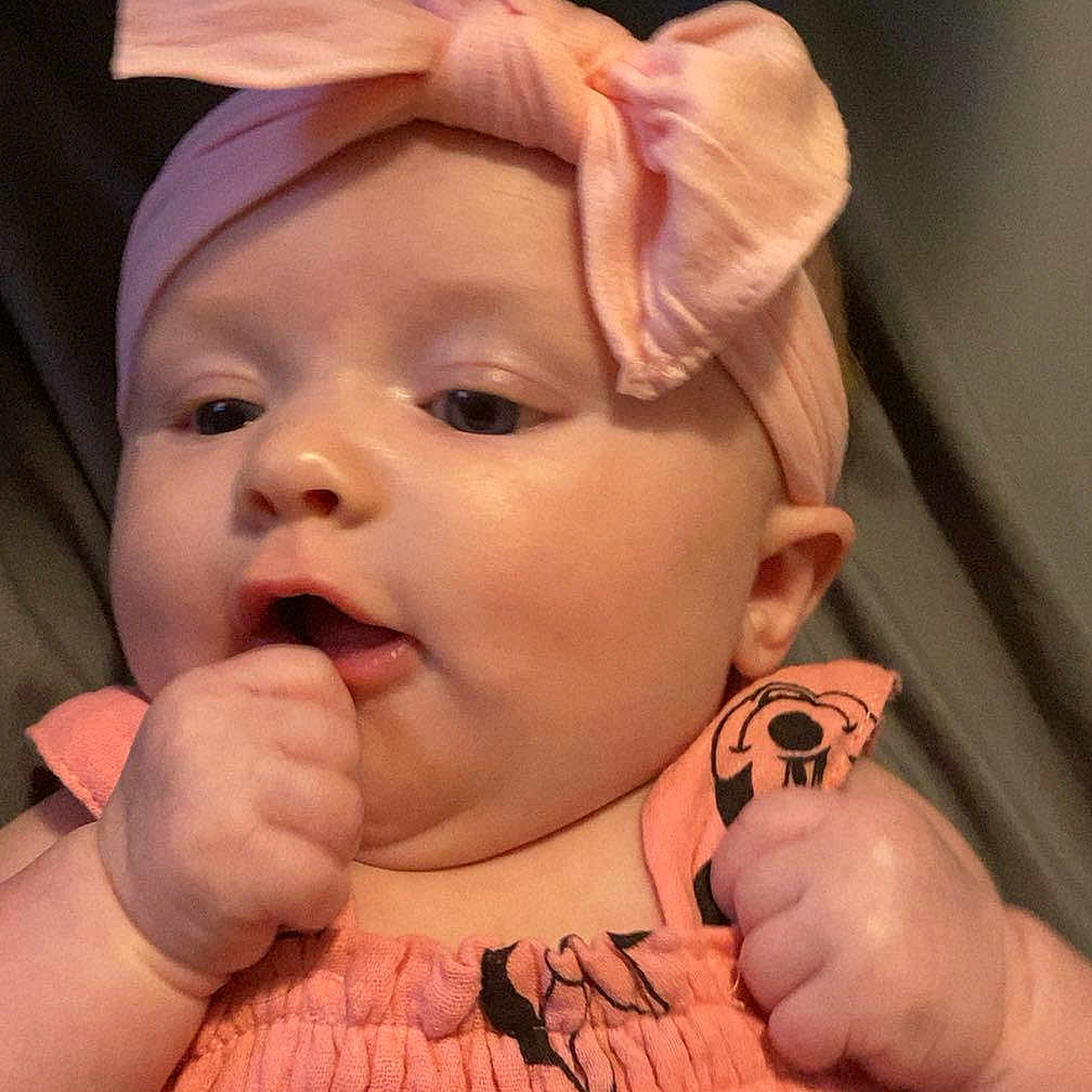 Navianna is registered to the contest to win money with this photo: baby, bow, cartoon_print, cheeks, child, closeup, clothing, curious, cute, dress, face, hand, headband, indoors, infant, person, pink, portrait, skin, soft_lighting