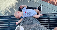 Wes is registered to the contest to win money with this photo: baby, bench, black_bench, casual, child, footwear, gray_clothing, lap, nap, onesie, outdoor, pacifier, palm_shadow, person, relaxing, shadow, sidewalk, sneakers, sunlight, tattoo