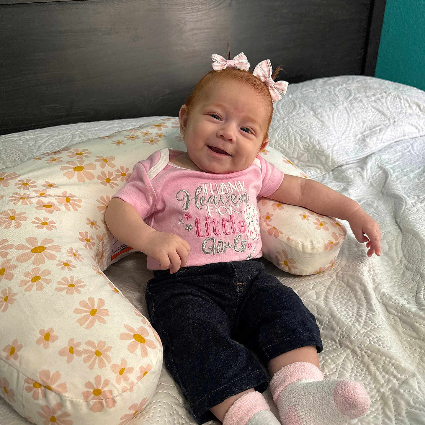 Haislyn joined the competition — help win amazing prizes! baby, infant, girl, smiling, pink_bows, pink_shirt, dark_pants, socks, floral_pillow, bed, quilted_bedspread, headboard, happy, cute, child, indoors, clothing, person, resting, posing