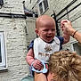 Vincent is registered to the contest to win money with this photo: baby, man, shoulders, bib, smile, happy, outdoor, courtyard, brick_wall, window, light_bulb, toy, hand_holding, person, child, hair, clothing, skin, face, playful