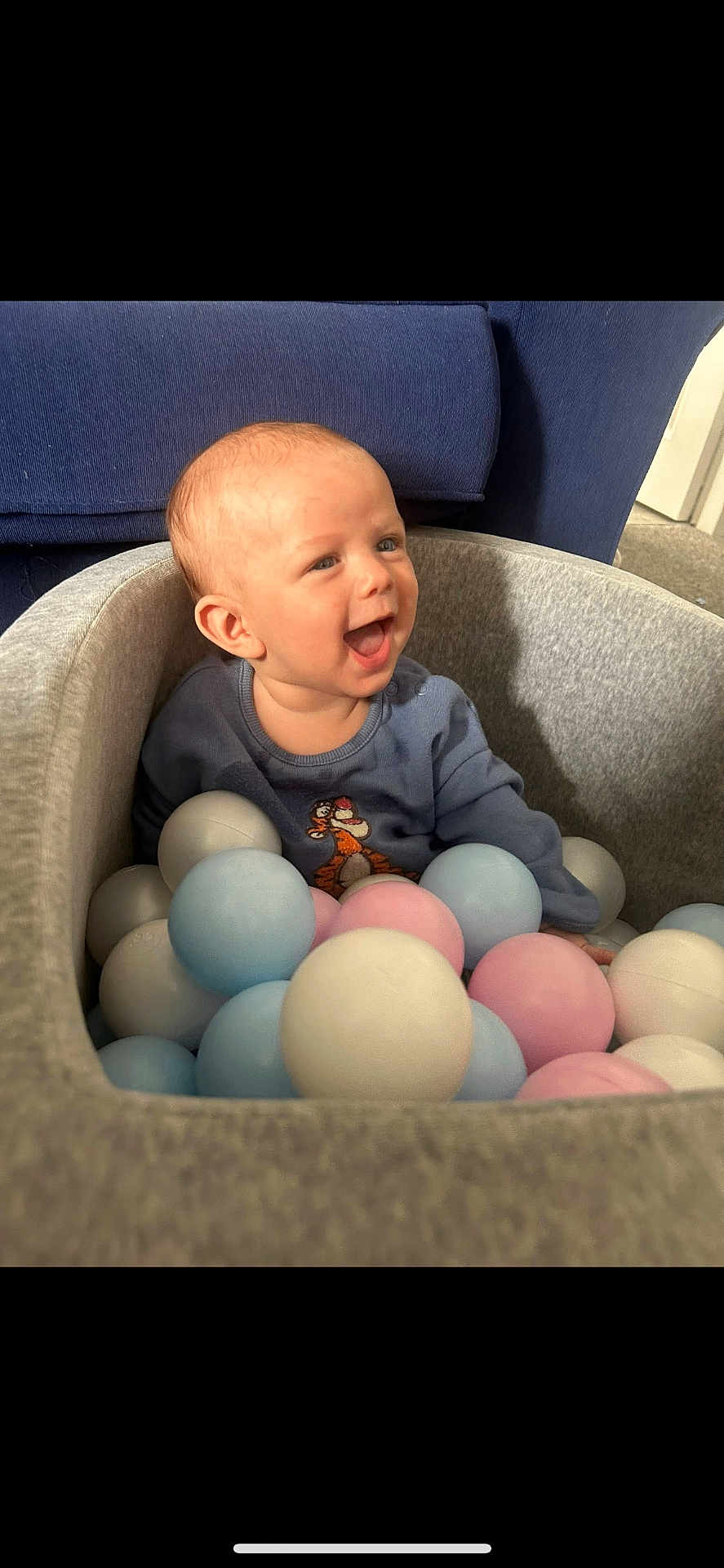Vincent is registered to the contest to win money with this photo: baby, child, ball_pit, blue_sweater, pastel_balls, smiling, happy, indoor, cozy, cute, toddler, playful, soft_lighting, seated, toy, person, face, joyful, blue_couch, grey_fabric