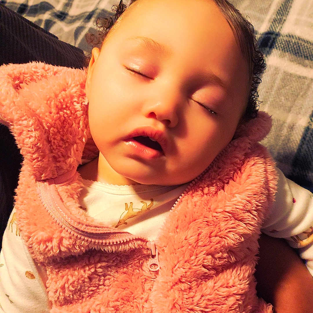 Maryah is registered to the contest to win money with this photo: baby, child, clothing, cozy, curly_hair, hand, indoor, infant, laundry_basket, peaceful, person, pink_vest, plaid_blanket, portrait, rest, sleep, sleeping, soft_texture, toddler, warm