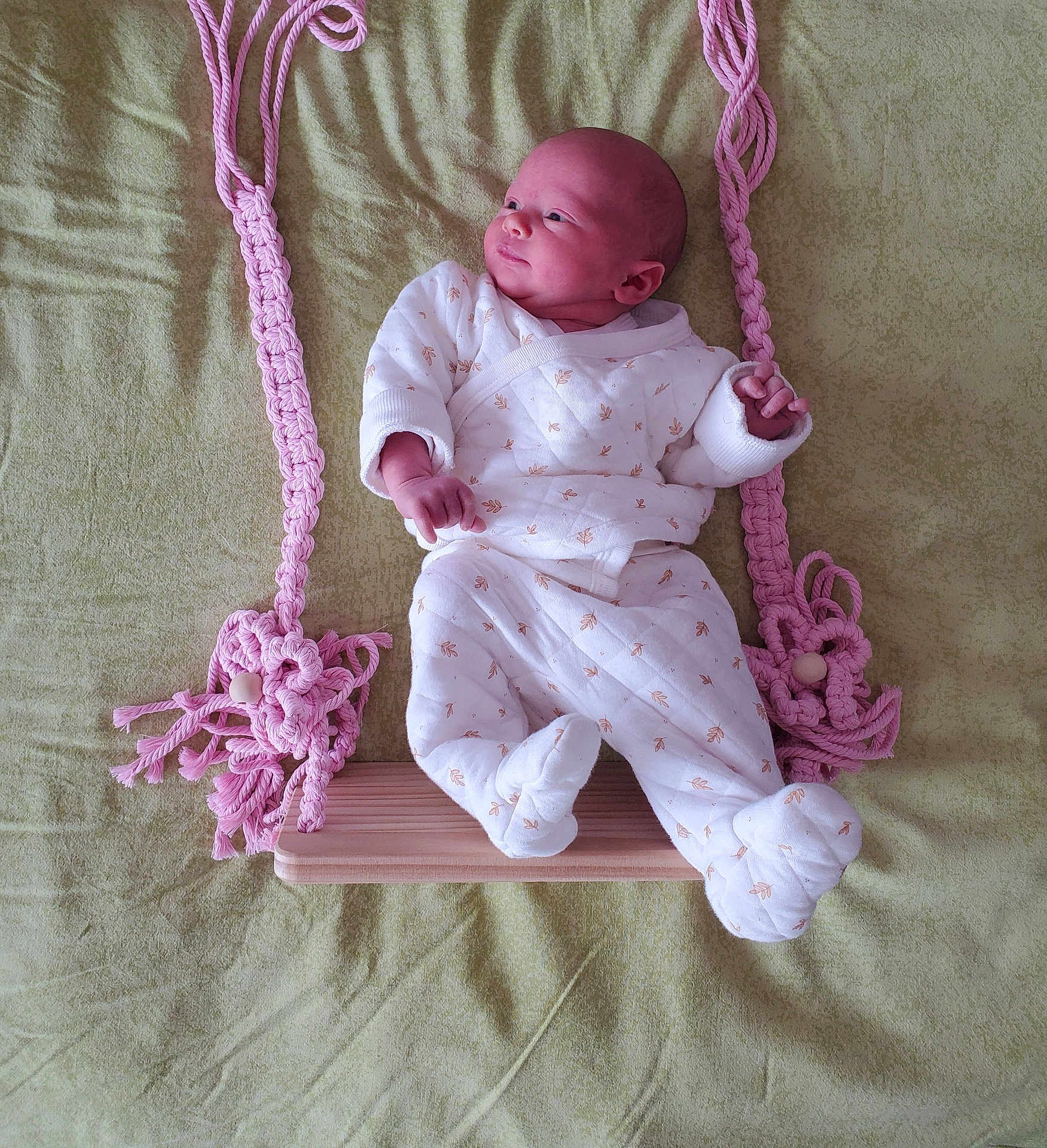 Maya a rejoint le concours — aidez-le/la à gagner de superbes lots ! baby, infant, swing, wooden_swing, pink_ropes, pajamas, white_clothing, patterned_clothing, lying_down, soft_background, green_background, indoors, cute, newborn, child, small_hands, small_feet, head, face, peaceful