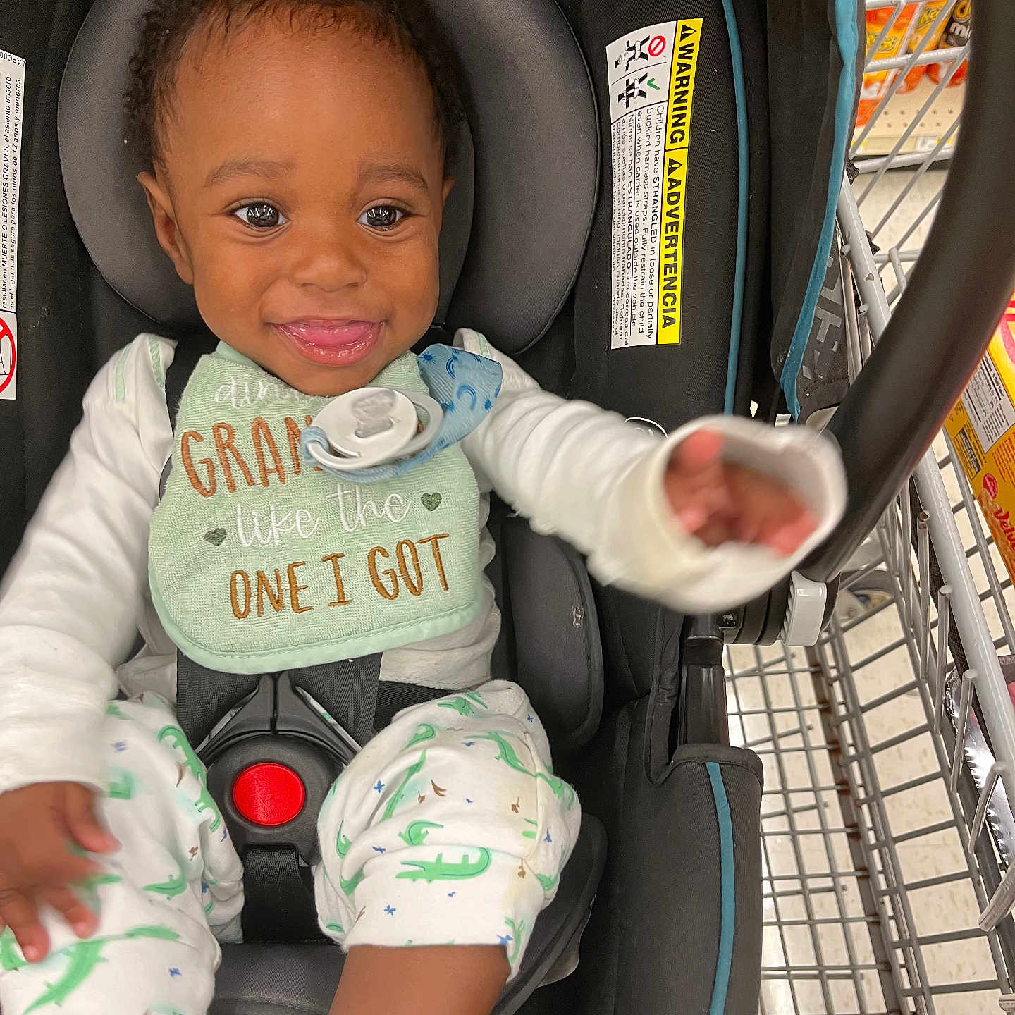 Dayton is registered to the contest to win money with this photo: aisle, baby, bib, car_seat, cart, child, clothing, cute, food_products, grocery_store, happy, indoor, infant, pacifier, person, reaching_hand, retail, seat_belt, shopping_cart, smiling