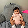 baby, infant, pink_onesie, socks, cushion, gray_cushion, bed, soft_texture, wall, indoor, person, child, cute, seated, portrait, smiling, looking_at_camera, skin, hair, small_person
